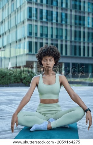 Relaxed dark skinned young woman practices yoga in city meditates and breathes deeply for stress relief poses in lotus pose on rubber mat wears sportsclothes improves inner balance serene state