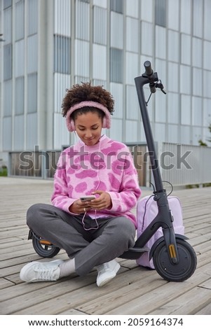 Relaxed teenage girl sits crossed legs near electric scooter rests after driving listens music via earphones and smartphone enjoys leisure time in city surfs social networks. People lifestyle hobby