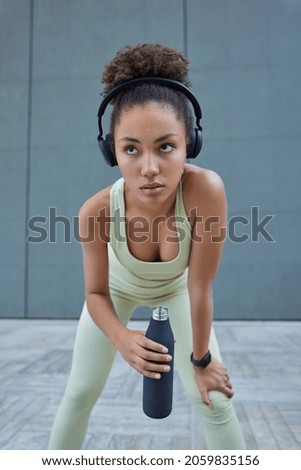 Tired sweaty sportswoman leans forward takes break holds bottle of water dressed in tracksuit looks cofident poses over grey wall listens music via headphones. Female model rests after cardio training
