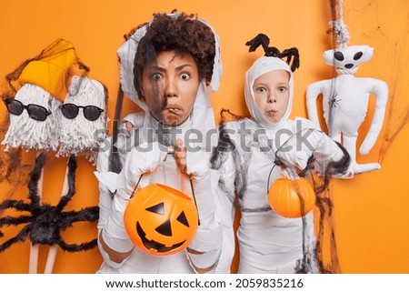 Horizontal shot of stupefied adult woman and girl wear ghost costumes hold carved pumpkins make jack o lanterns for halloween pose against orange background believe in mystery. Spooky creatures