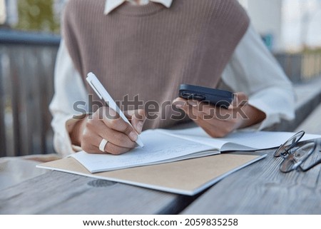 Unrecognizable woman writes down notes thesis for research report essay holds modern mobile phone holds pen makes list or plan engaged in time management activity sits at wooden table outdoors