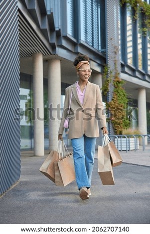 Full length shot of happy young female shopper in fashionable outfit returns from shopping mall carries many paper bags walks outside being in good mood makes purchases during big discounts.