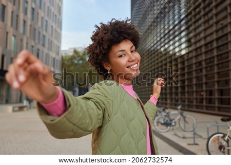 Cheerful carefree woman strolls in city and dances keeps arms sideways enjoys good day walks in downtown among modern city buildings wears jacket being in good mood. People leisure emotions.