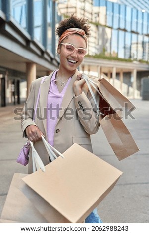 Beautiful cheerful woman enjoys big sales during shopping in Black Friday carries paper bags poses at street near mall wears pink sunglasses fashionable jacket smiles positively. Consumerism