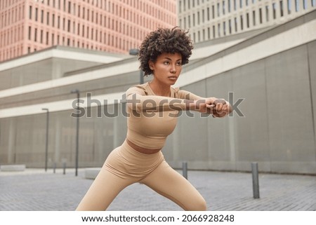 Fitness girl with curly hair does stretching exercises during sport training outdoors tries to improve health and physical form warms up for better flexibility poses at urbanity wears beige sportswear