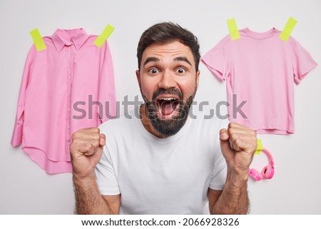 Photo of bearded young man yells with loud voice keeps mouth widely opened clenchesfists reacts on something awesome wears casual t shirt poses against white background with pink items of clothes