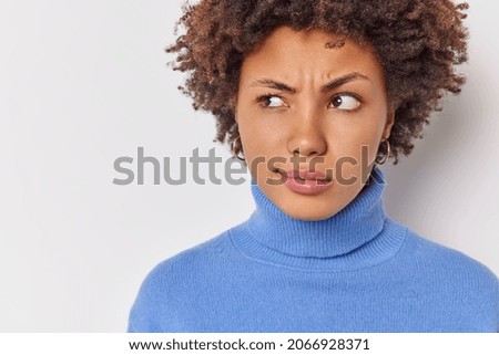 Close up shot of serious displeased curly haired woman looks attentively aside has suspicious expression dressed in casual blue turtleneck isolated over white background with blank space for advert