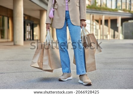 Unrecognizable female shopper in stylish jacket jeans and sneakers carries paper shopping bags made different successful purchases during Black Friday poses outside during daytime strolls in city