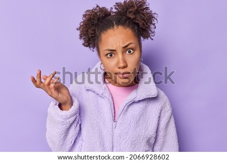Indignant puzzled millennial girl raises palm shrugs shoulders looks surprised at camera has clueless expression cannot decide dressed in artificial fur coat isolated over purple background.