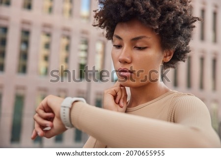 Healthy lifestyle concept. Curly haired woman checks pulse after jogging workout touches neck looks at watch dressed in sportswear stands outdoors against blurred background monitors her health