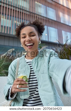 Happy optimistic girl with two hair buns dressed in jacket enjoys free time and walking in city holds bottle of detox drink makes selfie poses against modern building has fun during daytime.