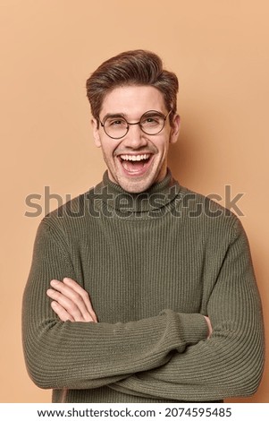 Cheerful confident handsome young European man with glad face expression keeps arms folded has positive talk with friend discuss something funny wears warm jumper poses against beige background.