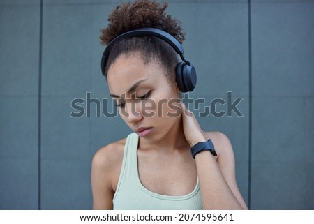 Sporty thoughtful woman with curly hair listens audio track in wireless headphones touches neck concentrated down takes break after workout poses against grey wall has intensive cardio training