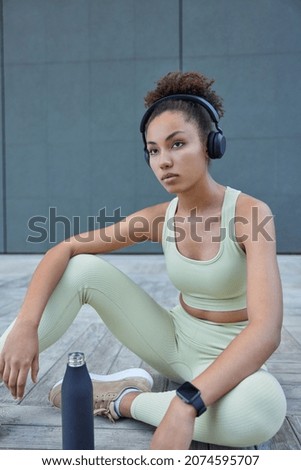 Thoughtful dark skinned sweaty sportswoman sits outdoors takes break after cardio training drinks water feels thirsty dressed in activewear listens music via headphones concentrated forward.