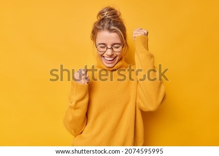 Yes finally success. Positive woman raises arms with clenched fists celebrates something has upbeat mood feels euphoric wears round spectacles and casual jumper poses against bright yellow background
