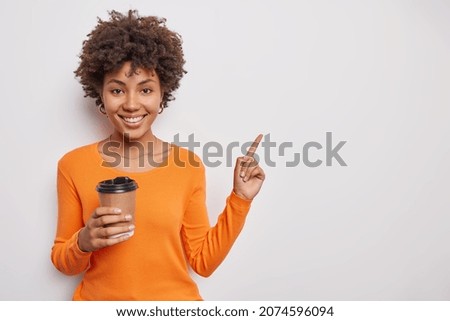 Positive good looking woman with curly hair wears casual orange jumper drinks takeaway coffee points at upper right corner shows copy space for your advertising content. Look at this please.