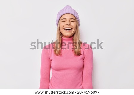 Joyful teenage girl laughs happily keeps eyes closed hears something positive wears knitted hat and pink turtleneck isolated over white background expresses sincere emotions. Happy woman poses indoor