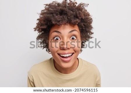 Close up shot of pretty cheerful woman gazes with happy surprised expression at camera smiles toothily wears casual t shirt isolated over white background looks with big interest at somethning amazing