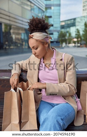 Fashionable young female shopper looks at products from herr shopping bags rests after purchasing poses at bench near modern mall in city feels tired wears fashionable clothes. Big sales concept