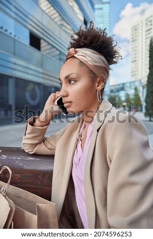 Vertical shot of thoughtful curly haired female shopper concentrated into distance talks via smartphone poses at bench with bags in modern big city rests after shopping day buys new trendy clothes