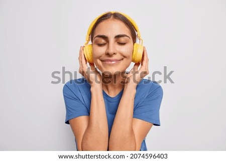Headshot of pleasant looking woman closes eyes from satisfaction enjoys favorite music in yellow headphones wears casual blue t shirt spends free time on favorite hobby poses against white background