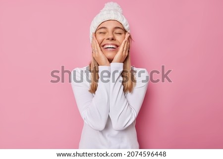 Photo of carefree cheerful girl keeps hands on cheeks smiles broadly closes eyes wears white knitted hat and turtleneck expresses sincere feelings isolated over pink background. Happy emotions