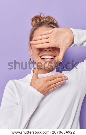 Smiling young woman hides face makes frame gesture takes photo keeps palm over eyes being in good mood wears casual white poloneck isolated over purple background captures moment pictures future