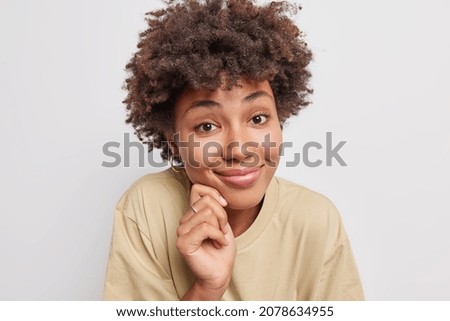 Portrait of lovely gentle woman with curly hair touches face and smiles pleasantly looks at camera has satisfied expression wears casual t shirt isolated over white background listens something good