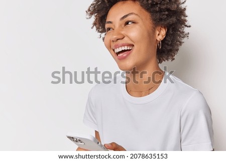 Indoor shot of happy curly haired woman smiles broadly receives pleasant notification on cellular has cheerful expression wears casual clothes isolated over white backgrund downloads new application