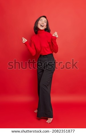 Full length shot of beautiful brunette woman dances carefree keeps hands raised wears turtleneck and black trousers isolated over vivid red background. Energetic carefree female model poses indoor