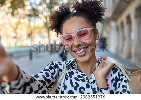 Positive carefree woman with two hair buns wears trendy pink sunglasses and blouse carries shopping bags poses outdoors during daytime. Smiling female shopper poses for making selfie in city