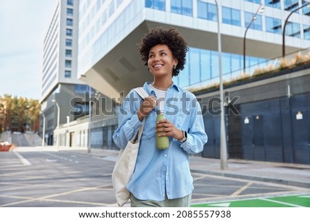 Waist up shot of happy young curly haired woman dressed in casual shirt carried fabric bag holds fresh water in bottle smiles positively strolls at city among glass skyscrapers enjoys spare time.