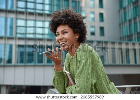 Happy curly haired woman dressed in green jumper holds notepad uses voice command recorder on smartphone translates words with application stands against blurred city background records message