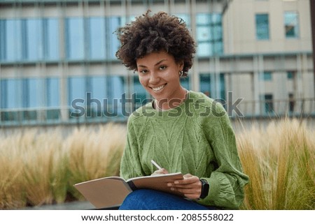Happy curly haired woman works on writing essay puts down ideas in notepad wears casual green jumper smiles positively at camera poses outdoors against blurred background creats text publication