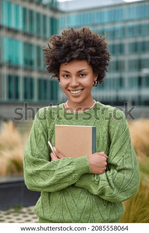 Vertical shot of happy young woman with curly hair holds notepad and pen makes notes what she observes around in city dressed in casual green jumper poses outdoors against blurred background