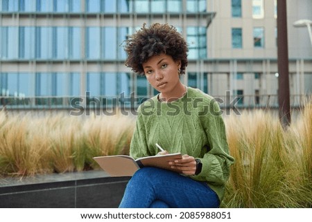 Horizontal shot of serious creative woman in jumper and jeans writes notes in notepad makes list to do poses outdoors against modern glass building puts down organization plan. Education concept