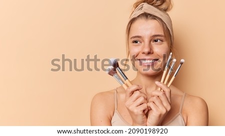 Makeup and beauty concept. Smiling young European woman with healthy glowing skin hods cosmetic brushes going to apply powder on face isolated over beige background blank copy space on left.