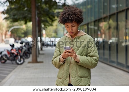 Photo of curly haired young woman in jacket uses mobile phone for chatting online strolls outdoors against blurred urban background holds modern device downloads application for finding route