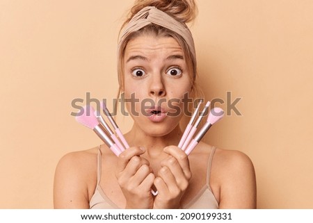 Shocked young European woman holds cosmetic brushes undergoes beauty routine uses new tools reacts on something astonishing wears headband and casual t shirt isolated over brown studio background