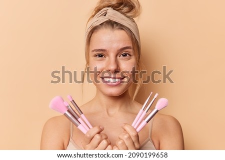 Beauty portrait of lovely pleased young woman smiles happily holds cosmetic brushes for applying makeup wears headband and casual t shirt looks directly at camera isolated over brown background