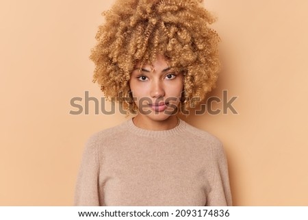 Portrait of serious confident sassy good looking woman with bushy curly hair looks directly at camera stands indoor against beige background wears casual jumper. Human face expressions concept