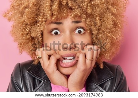 Headshot of worried curly haired woman bites finger nails looks nervously at camera afraids of something has anxious expression confused face isolated over pink background. Negative emotions