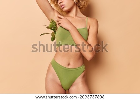 Unrecognizable faceless slim woman in green underwear demonstrates good physical shape perfect body and healthy skin poses against beige background. Sporty model wears cropped top and panties
