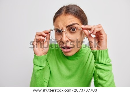 Serious brunette woman looks displeased stares through big spectacles has focused look tries to see something in distance wears green sweater isolated over white background.Hey what did you say