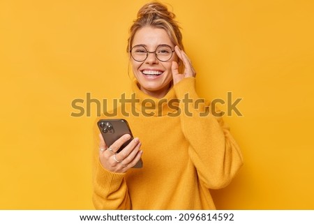 Positive woman smiles happily keeps hand on rim of spectacles feels cheerful wears comfortable sweater downloads cool application edits pics poses against yellow background. Technology concept