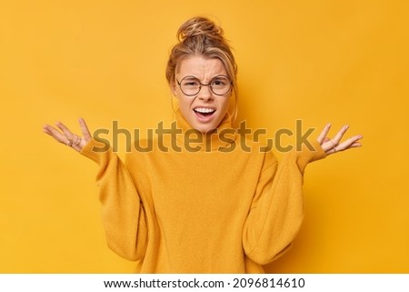 How could you. Annoyed indignant young woman spreads palms smirks face looks angrily at camera quarrels with someone feels puzzled wears spectacles and sweater isolated over yellow background Foto d'archivio © 
