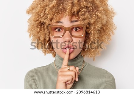 Mysterious curly haired young woman presses index finger over lips makes silence gesture asks to be quiet wears spectacles and turtleneck isolated over white background. Hush be quiet please