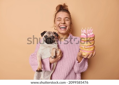 Happy woman poses with adorable oug dog and donuts going to blow candles celebrates pets birthday enjoys magic time wears knitted sweater isolated over beige background. Two best friends entertain