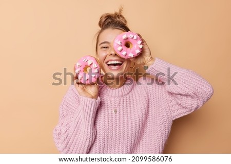 Happy joyful woman covers eye with delicious glazed doughnut laughs happily dressed in casual knitted sweater poses indoor against beige background has sweet tooth breaks diet. Junk food concept
