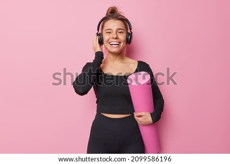 Happy sporty woman dressed in black sportswear carries rolled karemat poses at yoga studio going to have practice enjoys good sound in headphones isolated over pink background. Fitness concept